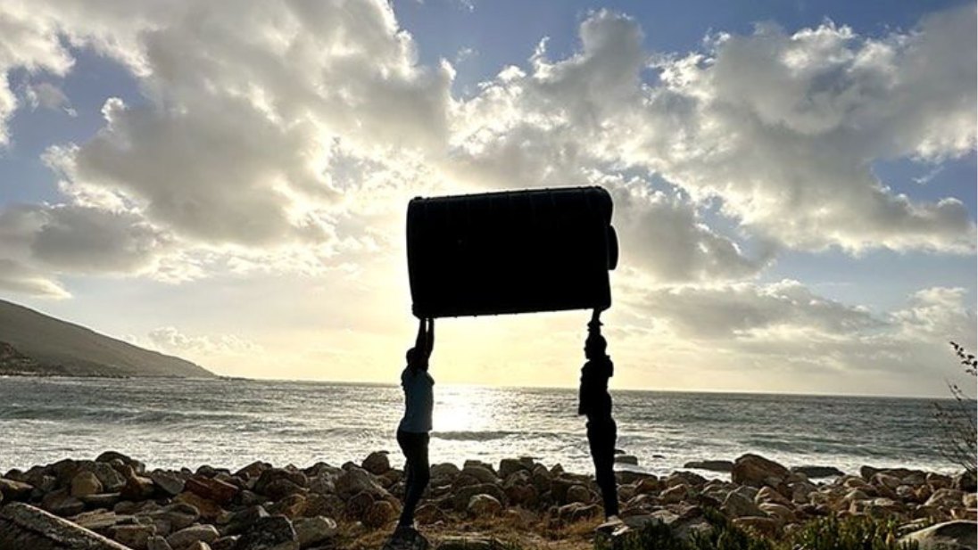 Two people lifting a water tank overhead on the Cape coast at sunset